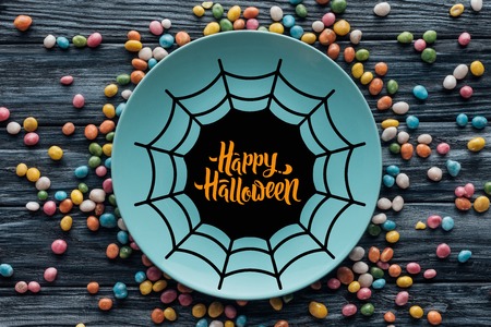 top view of plate with spider web and "happy halloween" lettering surrounded by colorful candies on wooden tableの写真素材