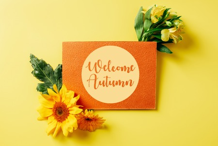 top view of orange card with "welcome autumn" lettering, sunflower, gerbera and lily flowers on yellow backdropの写真素材