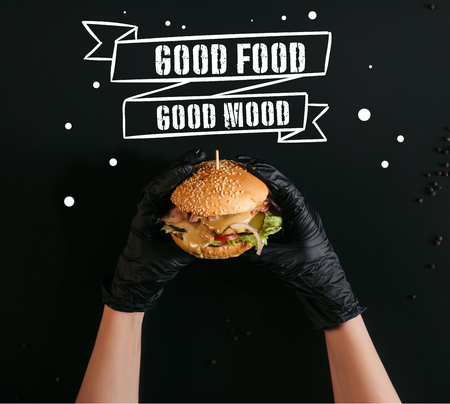 cropped shot of hands in gloves holding delicious burger with turkey, vegetables and caesar dressing on black with "good food - good mood" inspirationの写真素材