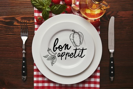top view of white plates with "bon appetit" lettering, cutlery and fresh basil with tomatoes and oil on wooden surfaceの写真素材