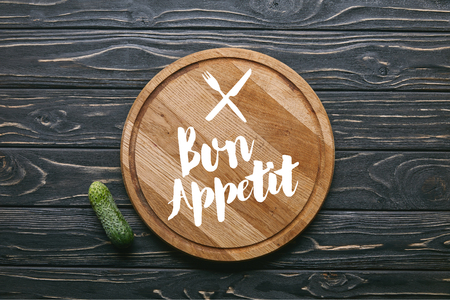 Cutting board with "bon appetit" lettering and cucumber on dark wooden tableの写真素材