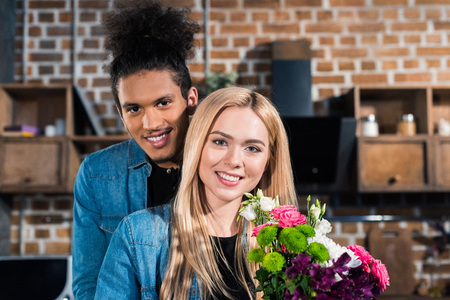 portrait of happy caucasian woman with bouquet of flowers and african american boyfriend near by in kitchen at homeの写真素材