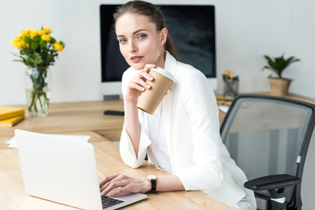 portrait of businesswoman with coffee to go at workplace with laptop in officeの写真素材