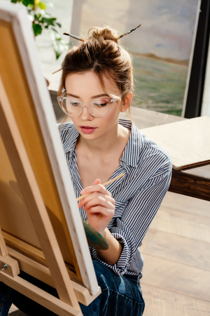 young stylish female artist in eyeglasses painting on easelの写真素材
