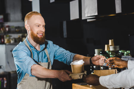 cropped shot of smiling bearded young barista giving paper bag and disposable coffee cups to african american buyer in cafeの写真素材