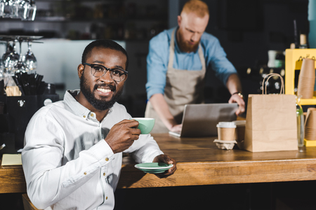 handsome young african american man drinking coffee and smiling at camera while bartender using laptop behindの写真素材