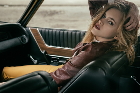 seductive young woman in leather jacket looking at camera while sitting in car