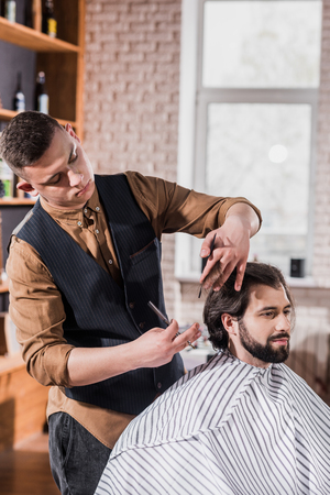 bearded young man covered with striped cloth getting haircut from professional barber at barbershopの写真素材