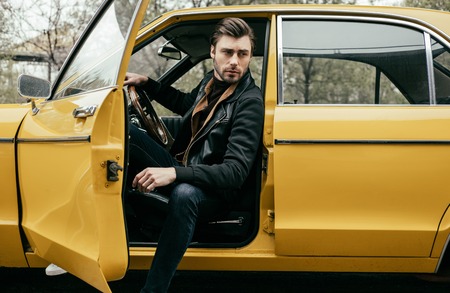 handsome stylish young man in leather jacket sitting in yellow classic car and looking away