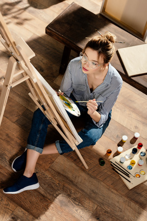 high angle view of stylish female artist in eyeglasses painting on easelの写真素材