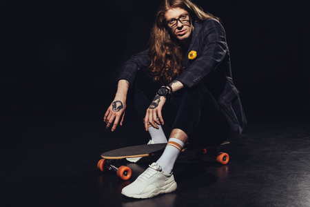 tattooed man with long hair sitting on longboard, on blackの写真素材