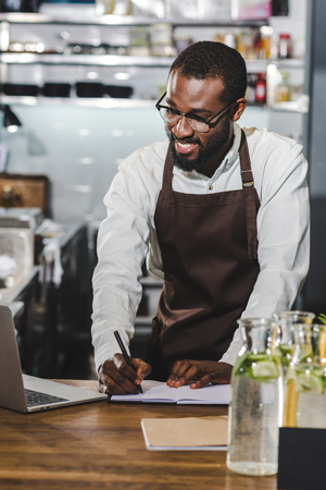 happy young african american barista taking notes and using laptop at coffee shopの写真素材