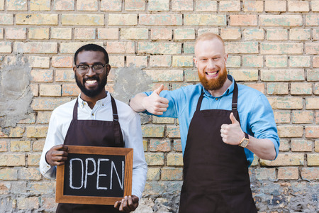 handsome multiethnic owners of coffee shop in aprons holding sign open and showing thumbs up,...