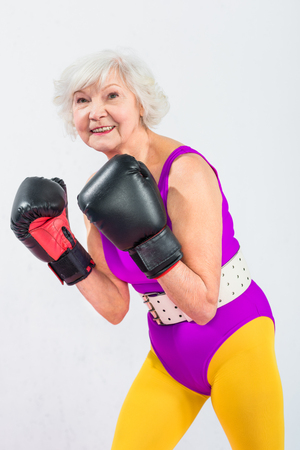 portrait of beautiful sporty senior lady in boxing gloves smiling at camera isolated on greyの写真素材
