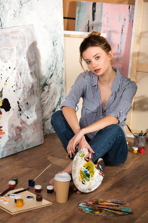 beautiful young artist sitting on floor and looking at camera in art studioの写真素材