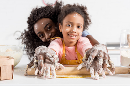 african american mother and smiling daughter rolling dough with rolling pin together on kitchenの写真素材