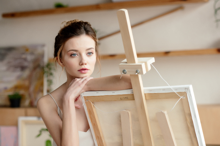 beautiful pensive young artist leaning at easel and looking awayの写真素材
