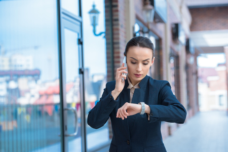 beautiful businesswoman checking time and talking by smartphone on streetの写真素材