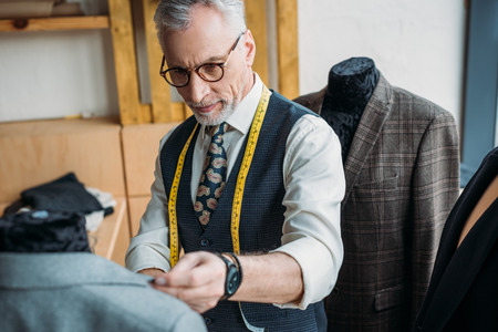 handsome mature tailor with measuring tape looking at jacket on mannequin at sewing workshopの写真素材
