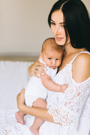 close-up shot of beautiful young mother with her cute baby sitting on bed at homeの写真素材