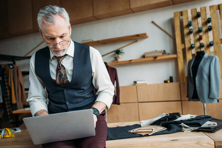 handsome mature tailor working with laptop while sitting on table at sewing workshopの写真素材