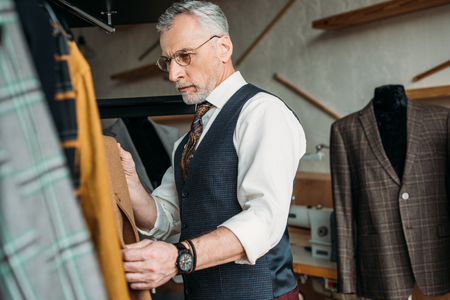 handsome mature tailor examining jackets at sewing workshopの写真素材