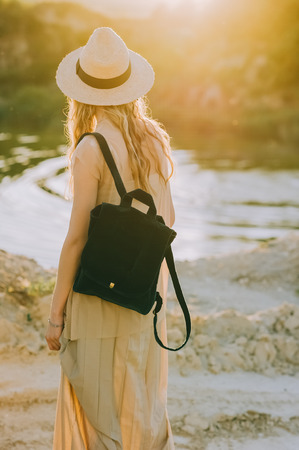 back view of blonde girl in hat with backpack standing near lake with sunlightの写真素材