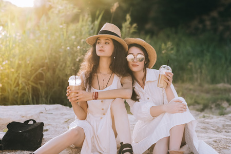 beautiful female friends in straw hats holding plastic cups with coffee latte and sitting on ground with sunlightの写真素材