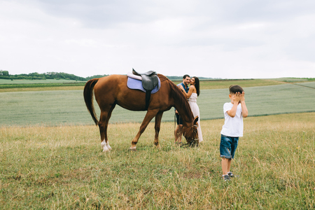 parents hugging near horse, son covering eyes on fieldの写真素材