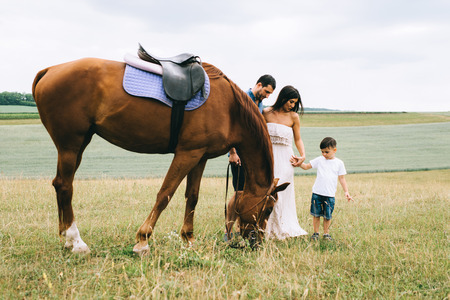 parents and son standing near beautiful brown horse on fieldの写真素材