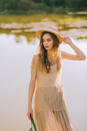 attractive blonde girl in stylish dress and straw hat posing near lakeの写真素材