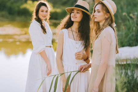happy attractive girls in straw hats standing near lakeの写真素材