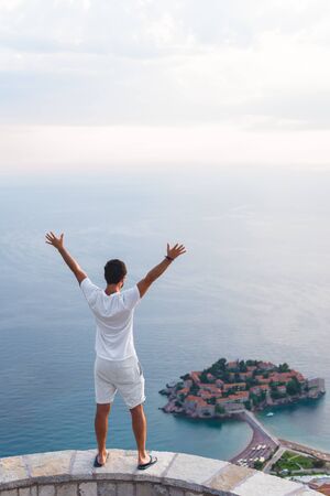 back view of man with raised hands looking at island of Sveti Stefan with hotel resort in Adriatic sea, Budva, Montenegroの写真素材