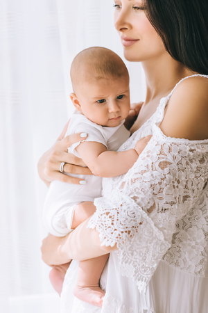 close-up portrait of young mother carrying her little baby in front of white curtainsの写真素材