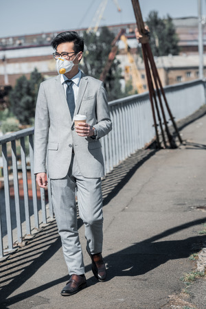 asian businessman in protective mask walking on bridge with coffee to go and looking away, air pollution conceptの写真素材