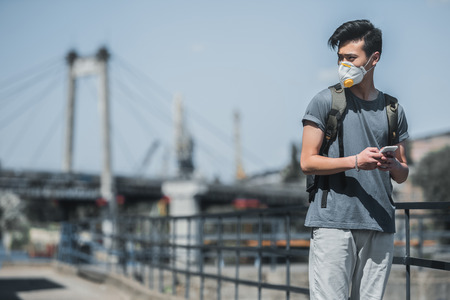asian teen in protective mask holding smartphone and looking away, air pollution conceptの写真素材