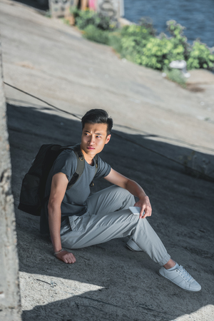 asian teenager sitting with bag and smartphone under bridge in cityの写真素材