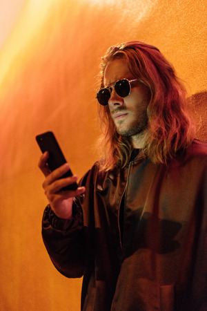 serious young man in leather jacket and sunglasses using smartphone on street at night under yellow...