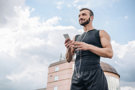low angle view of cheerful sportsman listening music with smartphone and looking away on roofの写真素材
