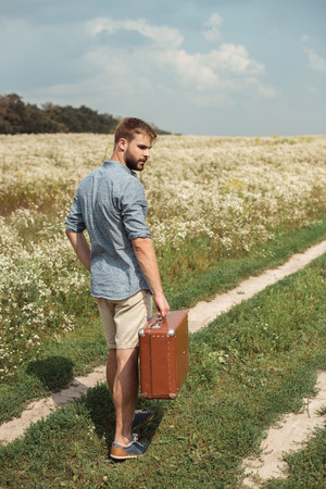 back view of man with retro suitcase standing in filed with wild flowers on summer dayの写真素材