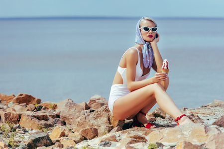 beautiful woman in vintage swimwear holding ice cream and posing on rocky beachの写真素材