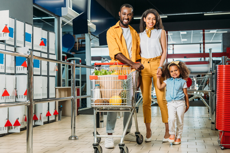 happy african american couple and their daughter standing near shopping trolley with food in supermarketの写真素材