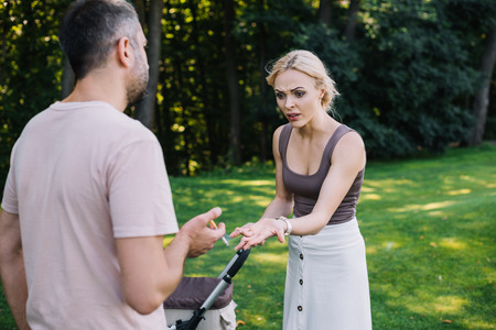 father smoking cigarette near baby carriage in park, mother pointing on cigaretteの写真素材