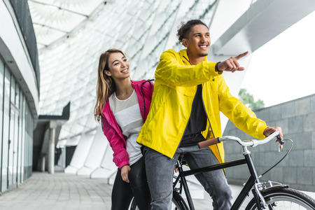 smiling mixed race man sitting on bicycle and pointing by finger to girlfriend at city street