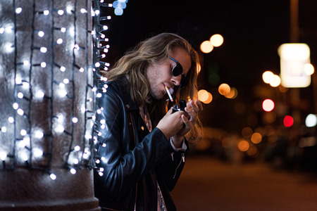 handsome young man in sunglasses and leather jacket smoking cigarette under garland on city street...