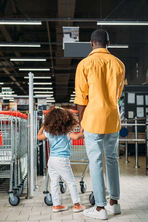 rear view of african american man standing with daughter near shopping trolley in supermarketの写真素材