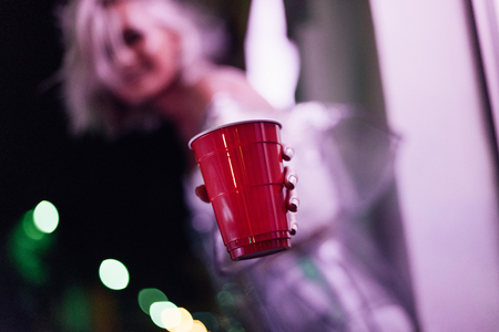 close-up shot of young woman holding red plastic cup on street at night under pink lightの写真素材