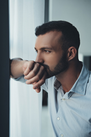 thoughtful man standing at window at homeの写真素材