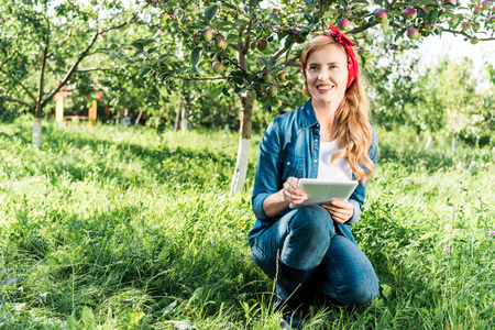 attractive farmer squatting and holding tablet in apple garden at farmの写真素材