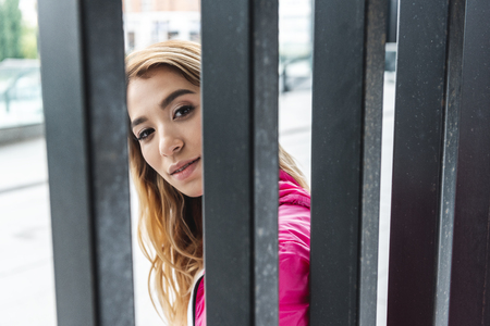 young stylish asian woman standing near metal fence and looking at camera at urban streetの写真素材